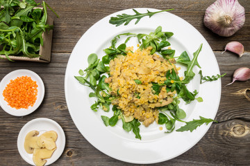 Red Lentils Dish with Mushrooms and Fresh Wild Rockets Leaves on White Plate. Raw Ingredients on Wooden Background.