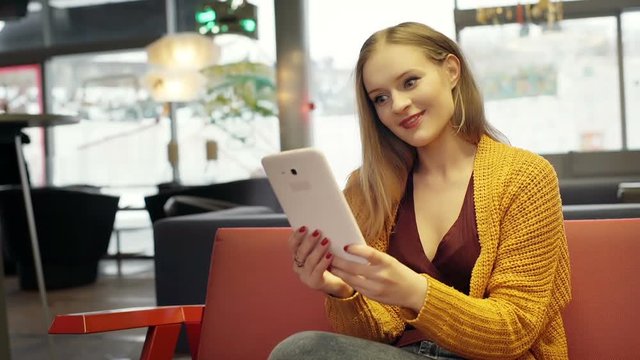 Pretty girl in yellow sweater having a videchat on tablet in the cafe

