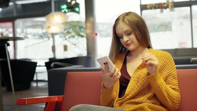 Happy girl sits down on the sofa in the stylish cafe and texting on smartphone
