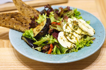 Vegetables salad of pepper, eggplant with mozzarella, pumpkin seeds and pesto sauce with brown bread, on a blue plate, wooden table