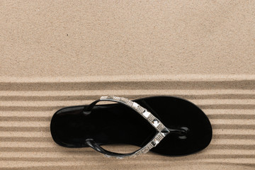 Trendy black flip flops decorated with rhinestones, standing on the beach sand.