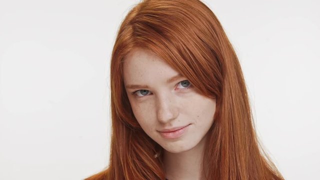 Shy charming ginger Caucasian teenage girl looking at camera lightly smiling. Close up footage on white background in slowmotion