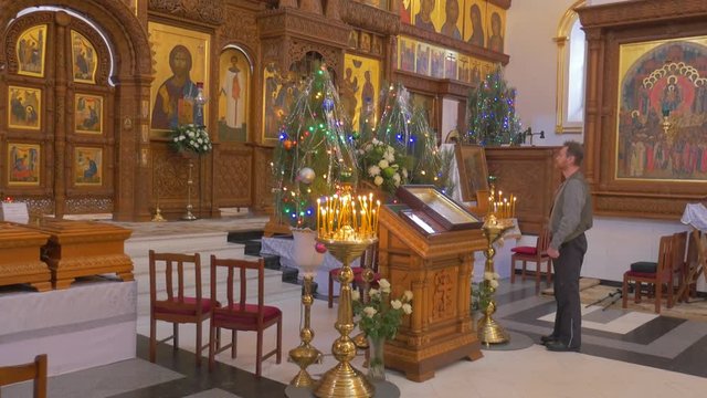 a Middle Aged Man Stands Before an Iconostasis, Which Looks Majestic, With a Lot of Lit Candles Before It, Fir Trees, and Golden Looking Icons