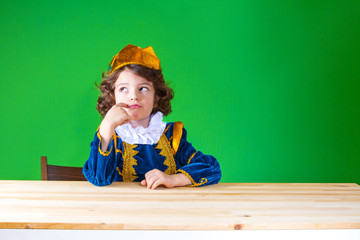 Sad curly Prince sitting at the table, looking thoughtfully up. Close-up. Green background.