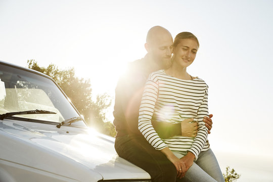 Young couple in love sitting on car bonnet at backlight