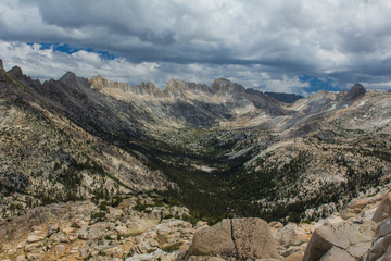 A threatening sky hangs over a wide and long forested alpine valley surrounded by jagged peaks