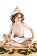 Cute curly-haired boy shirtless in a towel is sitting cross-legged with a wooden mug in hand, smiling, looking into the camera. Background bleached wood. © kravik93