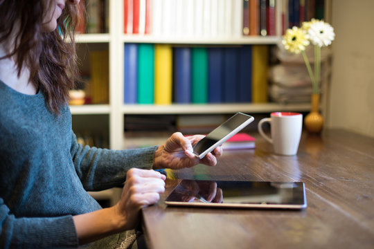 hands of adult brunette woman with green sweater using smartphone with screen tablet, multitasking, on wooden table with cup of coffee, library behind
