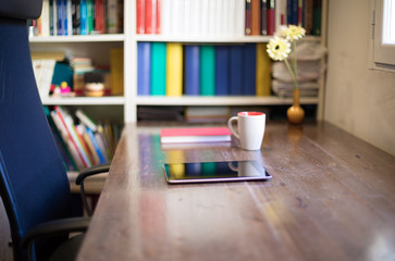 digital tablet, cup of coffee, book, vase with flowers on wooden table, black seat, and library in office next to window, nobody
