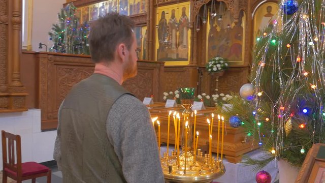 Christian Orthodox Cathedral on New Year`s Day, and a Bearded Man Praying Inside of it Before Some Saint Icon and a Decorated Fir Tree