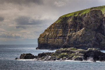 Lybster, Scotland - June 4, 2012: Huge brown-gray cliff with green meadow on top protects the south flank of Lybster harbor. It descends straight into the blue-gray sea under brown-gray skies