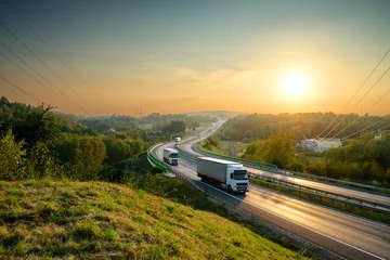 Fototapeten Weiße Lastwagen fahren auf der Autobahn, die sich bei Sonnenuntergang durch bewaldete Landschaft schlängelt. © am