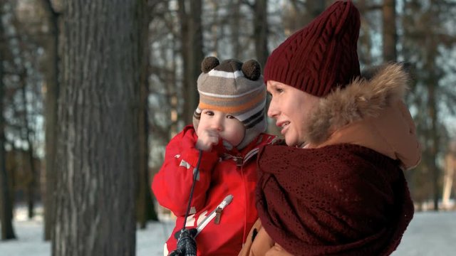 Young well dressed attractive beautiful mother with warm dressed little child in the park. She shows him something by hand. Little baby boy in winter warm jacket and funny bonnet or hood.