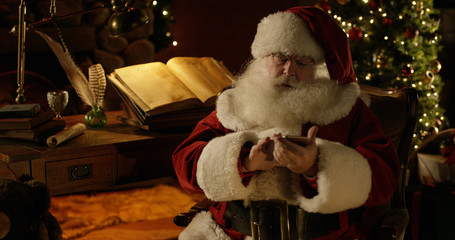 Santa Claus sits at his work bench in the North Pole, checking his smart phone.