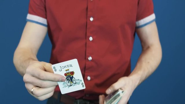 Close up magician in red shirt shuffle playing cards, show Jocker to camera. Blue background