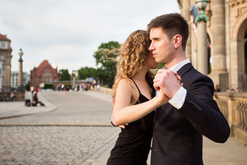two young people - a man in black suit and a woman wearing black dress - dancing tango outside; a musician is playing romantic songs; focus on the dancers hands