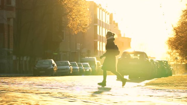 young travel woman in long black coat crossing paved road in the center of town with parked cars on both sides, road, trees and buildings are shining gold under the blind sunlight