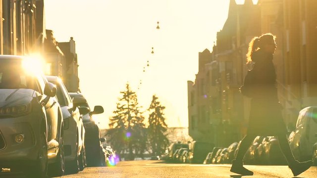 young travel woman in long black coat crossing paved road in the center of town with parked cars on both sides, road, trees and buildings are shining gold under the blind sunlight