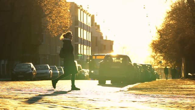 young travel woman in long black coat crossing paved road in the center of town with parked cars on both sides, road, trees and buildings are shining gold under the blind sunlight