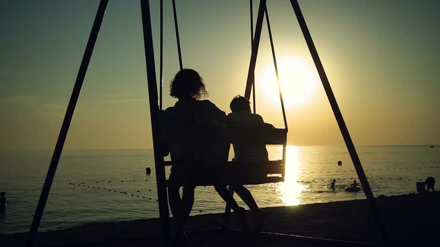 Silhouette of mother and her son swinging on a swing on a sea shore at sunset. HD