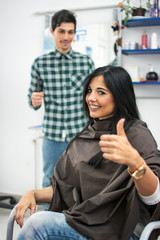 Young woman showing thumb up in hair salon. The hairdresser is standing behind her.
