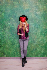 Obraz premium Closeup portrait girl with red balloons with heart shape on a green background. Vertical shot. Girl hipster in a black bowler hat holding a red inflatable heart and celebrate Valentine's day.