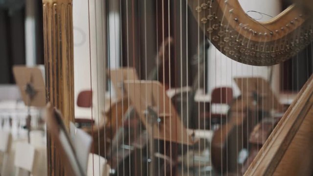 Close up dolly shot of harp strings with blurred music stands and cello on background