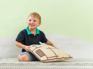 Cute blond kid playing with pillow, sitting cross-legged on a bed in the children's room. A broad...