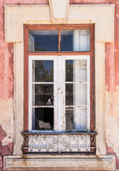Worn out window frame in Portugal. With broken glass and peeling paint. There are old ornate metal bars. The paint is pink and cream.