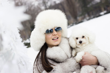Obraz premium Play time at the park. Woman with winter sunglasses playing with her dog. Young beautiful woman wearing a fur hat and a winter coat.
