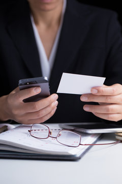 A business woman in black suit holding white blank business card in her hand with mobile phone in the other hand on a working desk.