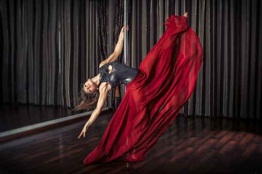 Young pole dancer girl in mask and red skirt sitting indoor near the pole on curtain background