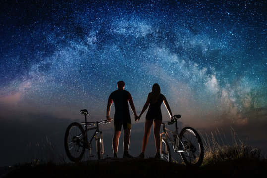 Rear view of silhouette couple cyclists holding hands enjoying night sky with lots of stars. Bikers with mountain bicycles on the hill. Night landscape with colorful Milky Way