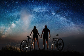 Obraz premium Rear view of silhouette couple cyclists holding hands enjoying night starry sky. Bikers with mountain bicycles on the hill. Night landscape with colorful Milky Way