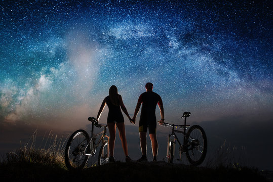 Rear view of silhouette couple cyclists holding hands enjoying night starry sky. Bikers with mountain bicycles on the hill. Night landscape with colorful Milky Way