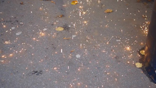 Sparks from the welding are falling down on the asphalt. A large piece of sizzling  hot metal hits the ground and shatters multitude of sparks, like water droplets scatters around