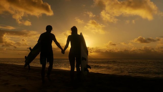 Surfer couple in silhouette holding long surfboards at sunset on tropical beach