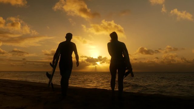Surfer couple in silhouette holding long surfboards at sunset on tropical beach