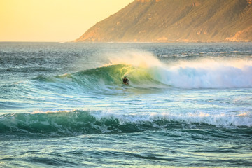 Noordhoek Beach at sunset. Surfing in Cape Town, South Africa. Surfer tries to take a big wave. Atlantic coast in Table Mountain National Park. Extreme sports leisure concept.
