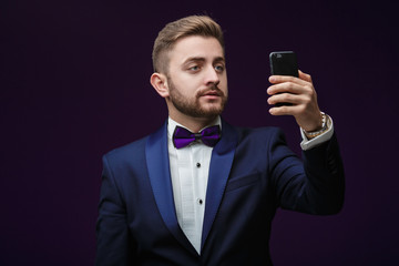 Naklejka premium handsome man in tuxedo and bow tie makes selfie against dark background. Fashionable, festive clothing