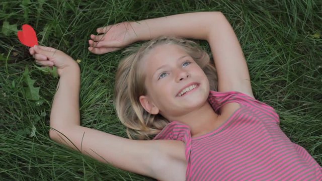 Happy smiling young girl lying on grass with arms outstretched having fun holding red paper heart in her hand. Love nature or happiness childhood concept