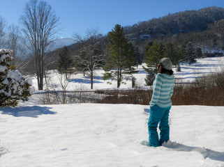 Woman standing on snow at a hill looking and admiring the winter trees mountains and Lake view wearing hat sweater and ski pants