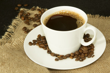White cup of coffee with coffee beans on saucer on linen cloth