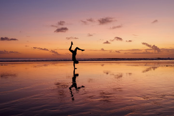 A silhouette of a girl and her reflection doing a handstand on a beach during sunset.