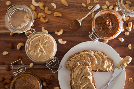 Jars of almond, cashew, and peanut butters sitting in sequence with their respective nuts lying around them