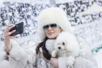 Beautiful woman taking a selfie with her dog outdoors. Winter time. Woman wearing a white fur coat. Christmas decorations in the background.