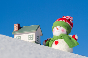The snowman stands next to the house on a snow slope. Blue sky. Winter background.