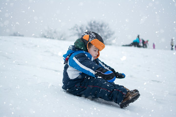 Cute little boy sledding down a hill on cold snowy on winter day.