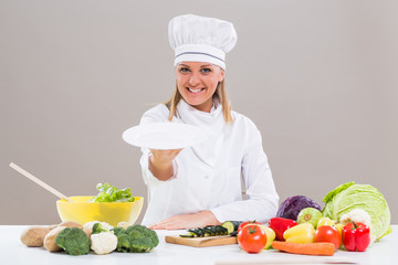 Portrait of cheerful female chef sitting at the table and showing empty plate while making healthy meal.
