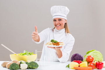 Cheerful female chef is sitting at the table with bunch of vegetable ,showing prepared meal and thumb up.
